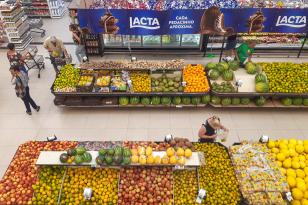 Ambiente de supermercado com prateleiras e bancas com frutas, legumes e produtos industrializados, além de alguns clientes realizando compras. 